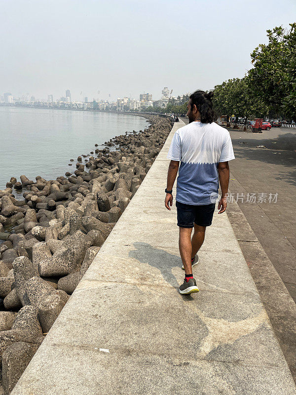 一个人走在海堤上的特写，孟买，印度，混凝土防御，海岸管理，防止侵蚀，海水，c形长廊，装饰艺术建筑和摩天大楼排列在Girgaon Chowpatty公共海滩的后视图
